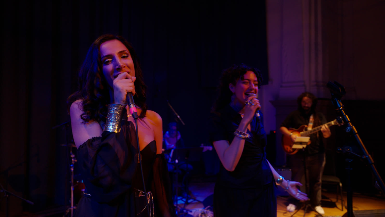 Sura singing a duet with a second vocalist in blue-violet light